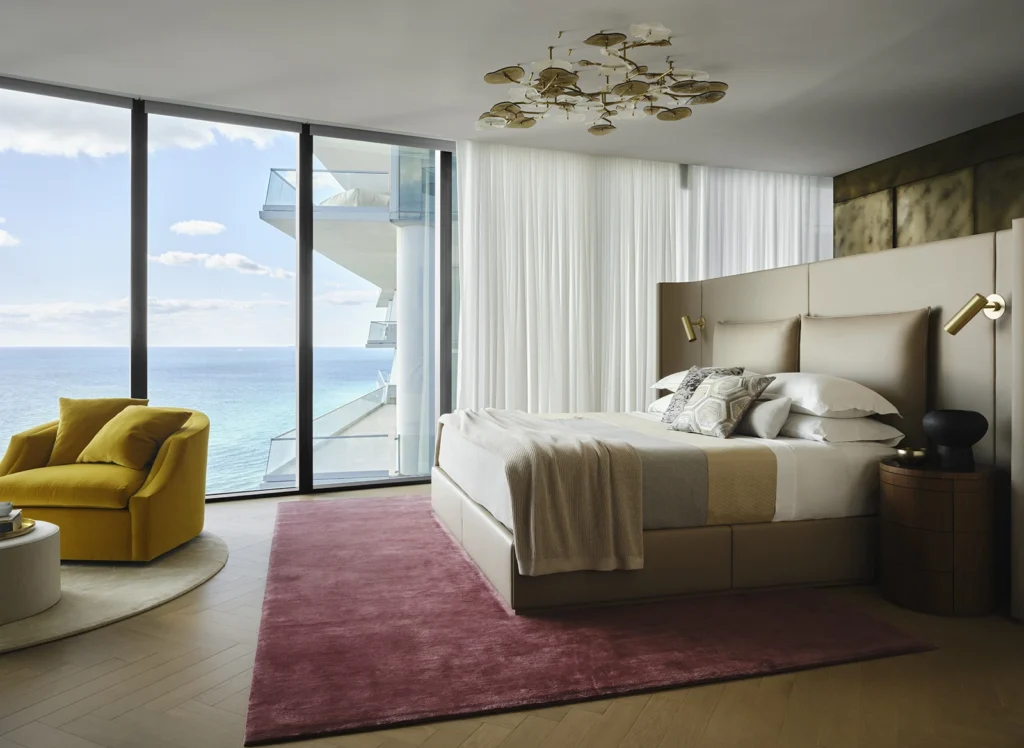 Modern bedroom design featuring a beige upholstered bed with decorative pillows, a yellow accent chair, a round coffee table, and large windows showcasing an ocean view, emphasizing luxury and craftsmanship in interior decor.